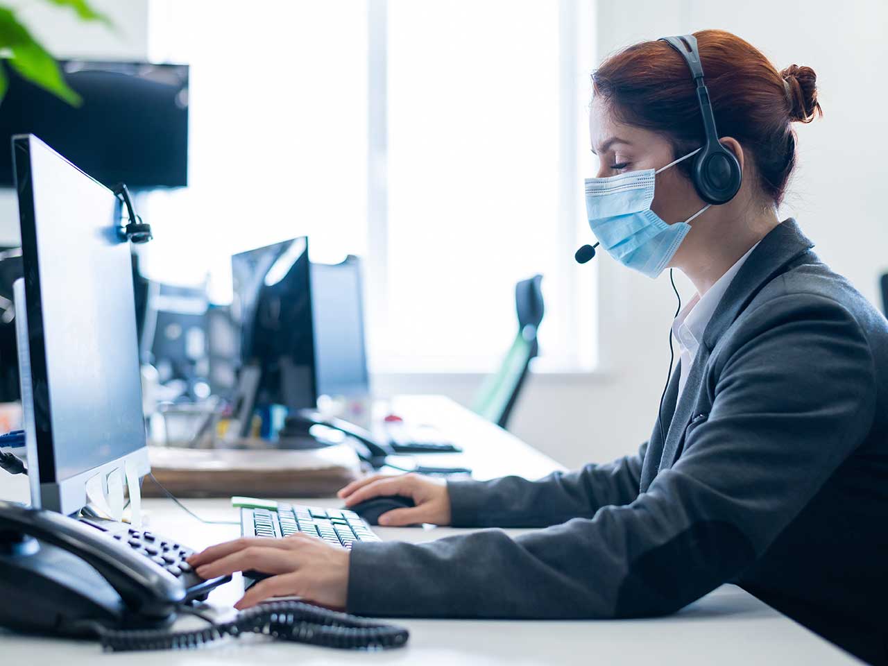 woman wearing surgical mask in office setting at desk with computer and phone