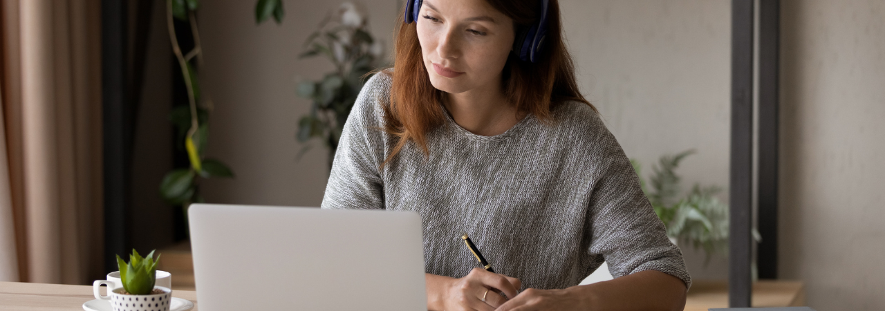 woman wearing headphones, taking notes and studying on laptop