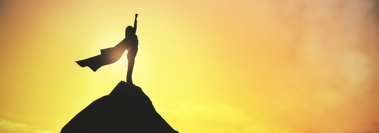 silhouette of a superhero with a cape standing atop a mountain with the sunset in the background