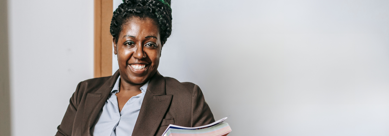 A teacher with a joyful expression, wearing a brown suit, holding a stack of books, and standing in front of a whiteboard