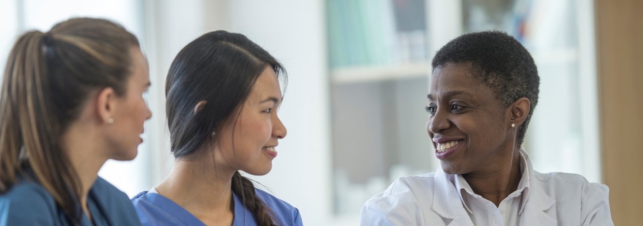 Three healthcare professionals, one in a white coat and two in blue scrubs, engaged in a discussion in a bright office setting.