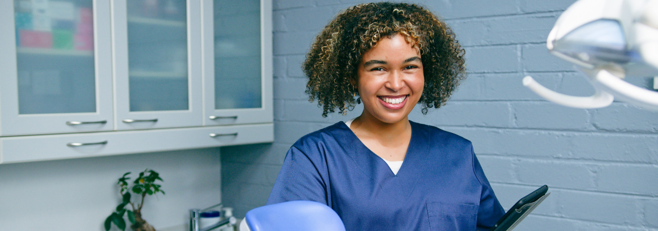 A dental assistant smiling and holding a tablet in an exam room
