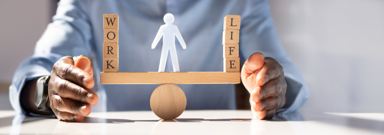 Person holding a balanced wooden scale with blocks spelling ‘WORK’ on one side and ‘LIFE’ on the other.