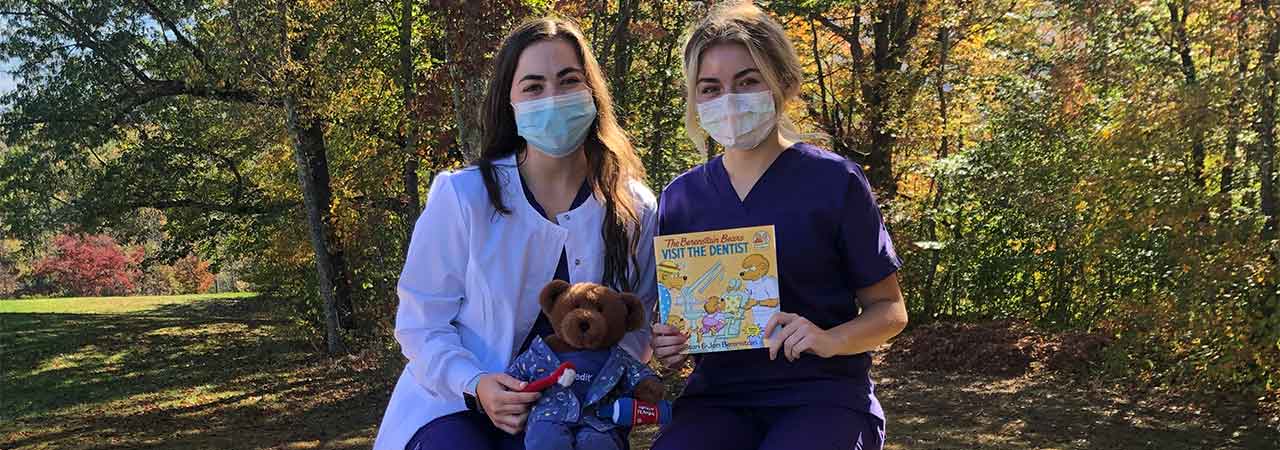 Dental assistants wearing masks outdoors with teddy bear and dental related children’s book